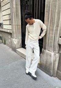 Kremowy sweter dziergany z dekoltem sznurowanym, zestawiony z białymi spodniami o szerokich nogawkach i masywnymi beżowymi butami. Miejska sceneria z kamienną ścianą.