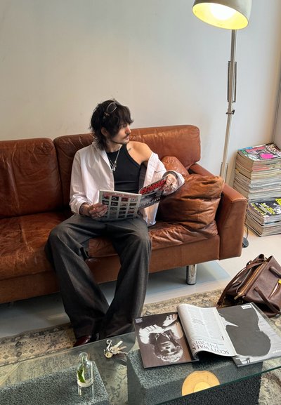 Sofá de cuero marrón con una superficie texturizada. Persona con camiseta de tirantes negra y camisa a rayas leyendo una revista. Mesa de café de vidrio con revistas y llaves.