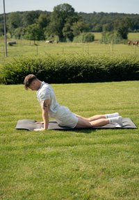 Uma pessoa executa uma pose de yoga num tapete preto sobre a relva. Ela veste uma camisa azul clara e calções brancos, com meias brancas.