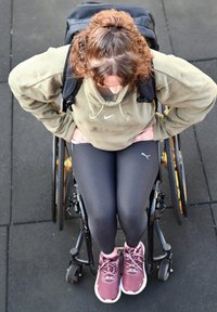 Schwarzer Rollstuhl mit gelben Akzenten. Person trägt einen grünen Kapuzenpulli und schwarze Leggings mit einem Puma-Logo. Kastanienbraune Sportschuhe mit weißen Sohlen.