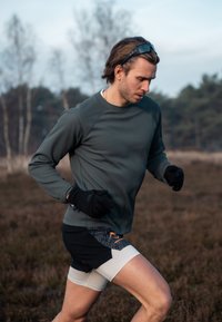 Un homme fait du jogging en portant un t-shirt gris texturé à manches longues et des gants noirs, assortis à un short noir et gris avec un accent orange.