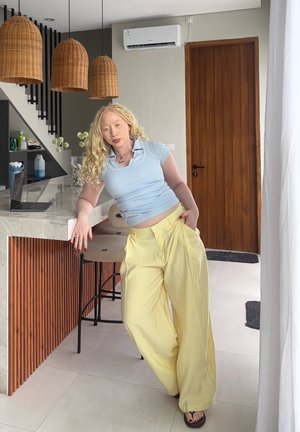 Mujer de piel clara y cabello rubio rizado se recuesta en la encimera de la cocina, vestida con un top azul claro y pantalones amarillos de pierna ancha en un entorno doméstico moderno.