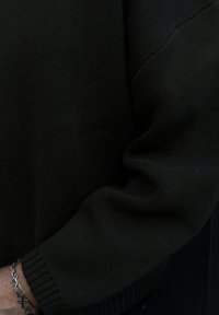 Pull en tricot vert foncé avec une surface texturée, doté d'un poignet côtelé. Le tissu semble doux et flexible, avec une coupe ajustée.