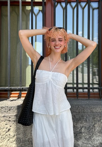 Top strapless de lino blanco con detalles fruncidos, combinado con una falda blanca fluida. Un bolso de paja negro descansa sobre un hombro. Ambiente de luz diurna brillante.