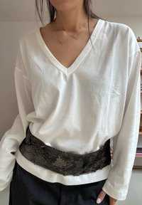 Haut blanc à manches longues avec col en V, coupe ample, associé à une ceinture en dentelle noire texturée avec un motif floral. Pantalon noir visible.