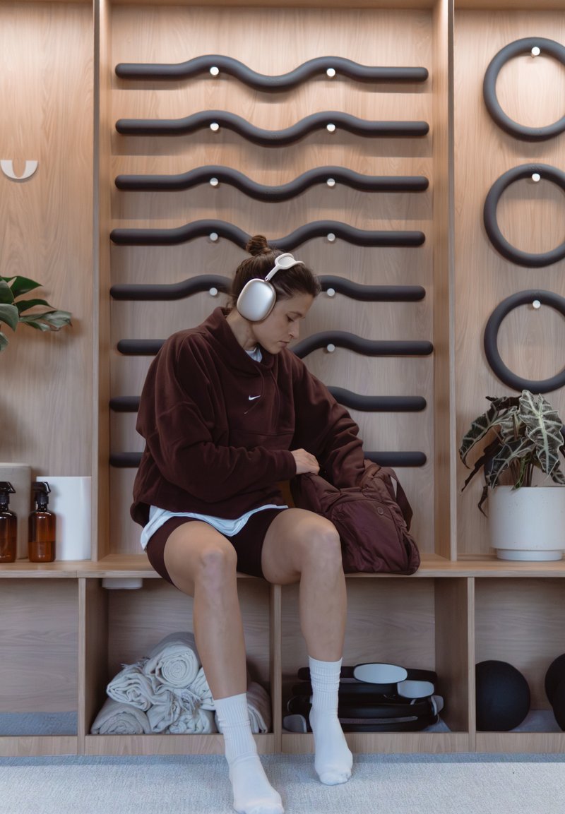 Femme portant un casque audio et une tenue de sport assise sur un banc en bois, cherchant dans son sac dans un studio de fitness ou de yoga moderne.