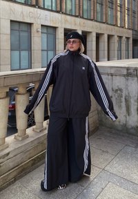 Μαύρο αθλητικό κοστούμι με λευκές ρίγες στους μανίκους και στα πόδια, που περιλαμβάνει ζακέτα με φερμουάρ και παντελόνια σε φαρδύ σχέδιο, συνδυασμένο με μια μαύρη μπερέ.