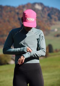 Osoba v růžové kšiltovce a modrém dlouhém rukávu si upravuje fitness hodinky, stojí venku s podzimními kopci v pozadí.