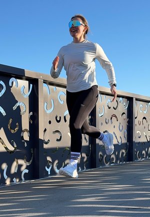 Camisa blanca de manga larga, leggins negros y zapatillas de correr blancas con detalles azules; usando gafas de sol reflectantes; corriendo en un puente texturizado.