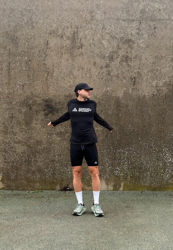 Un homme vêtu de vêtements de sport noirs se tient les bras écartés devant un mur en béton patiné par le temps. Il porte des chaussettes blanches et des chaussures de running.