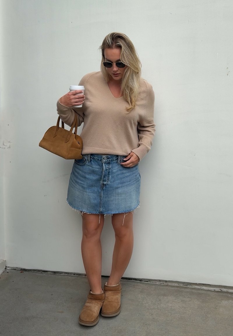 Donna con occhiali da sole, maglione beige, gonna di jeans, stivaletti marroni, che tiene una borsa marrone e una tazza di caffè bianca davanti a un muro grigio semplice.