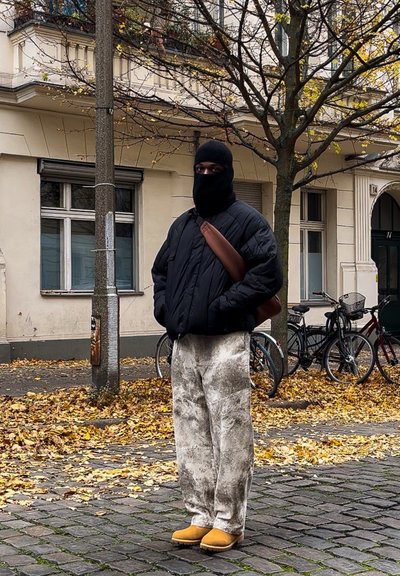 Chaqueta negra inflada, pantalones anchos de estampado blanco y zuecos amarillos. De pie sobre adoquines con bicicletas y hojas de otoño de fondo.