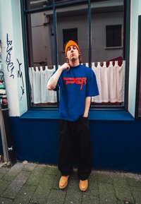 Modrý oversized tričko s červeným potiskem, černé široké kalhoty a hnědé dřeváky. Oranžový čepice dotváří ležérní vzhled.