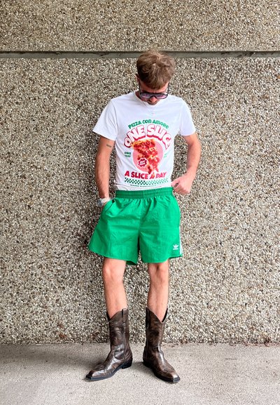 Camiseta blanca con un gráfico colorido de pizza y texto, pantalones cortos verdes y botas de vaquero marrones. Fondo texturizado liso.