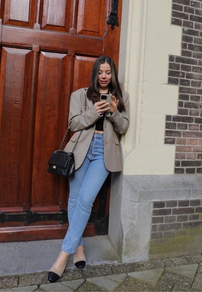 Blazer beige, top corto negro, jeans azules claros, zapatos negros y beige, y un bolso acolchado negro. De pie junto a una puerta de madera.