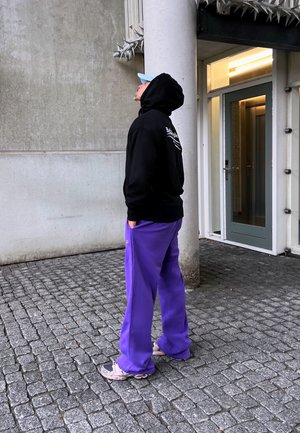 Persona con sudadera negra, pantalones morados y zapatillas en pie sobre un suelo de adoquines, mirando hacia arriba al lado de un pilar de concreto y la entrada de un edificio.