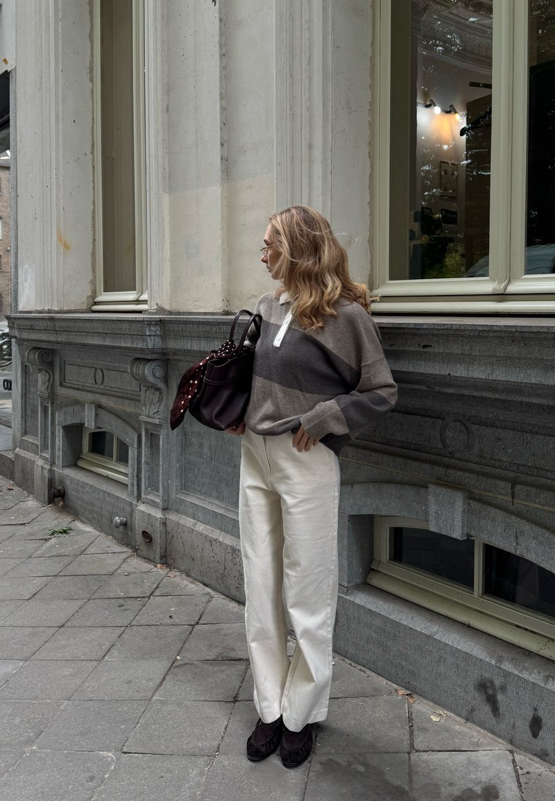 Maglione a righe grigio e beige oversize con colletto, pantaloni bianchi e scarpe scure. Borsa nera con sciarpa a pois. Ambientazione urbana.
