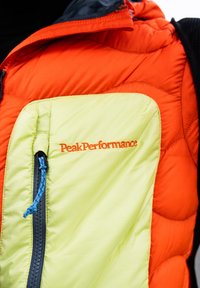 Orangene und hellgelbe gepolsterte Jacke mit blaurem Reißverschluss und "Peak Performance" auf der Brusttasche gestickt.