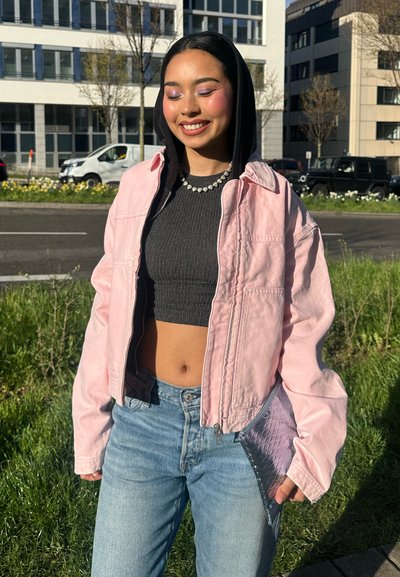 Joven sonriente con los ojos cerrados, lleva chaqueta rosa sobre sudadera negra con capucha, camiseta corta, vaqueros azules y sostiene un bolso de mano plateado al aire libre.