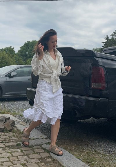 Camisa blanca de botones con rayas verticales, atada a la cintura; falda blanca con volantes en capas; sandalias marrones; gafas de sol en la mano.