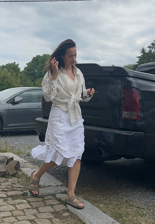 Witte overhemd met verticale strepen, geknoopt in de taille; gelaagde witte rok met ruffles; bruine sandalen; zonnebril in de hand.