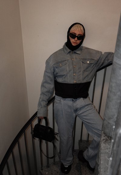 Chaqueta de mezclilla con un bajo negro corto, jeans de mezclilla azul claros, mascarilla negra y gafas de sol. Sostiene un pequeño bolso negro, de pie en una escalera de caracol.
