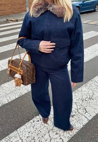 Personne portant une veste en jean bleu foncé avec col en fourrure et pantalon assorti, tenant un sac à main marron monogrammé avec un porte-clés ours en peluche sur un passage pour piétons.