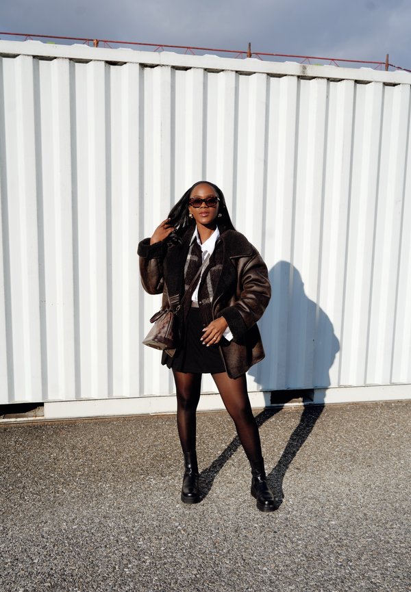 Cappotto in shearling marrone con dettagli neri, camicia bianca, gonna nera e collant. Stivaletti neri e una piccola borsa a tracolla marrone.