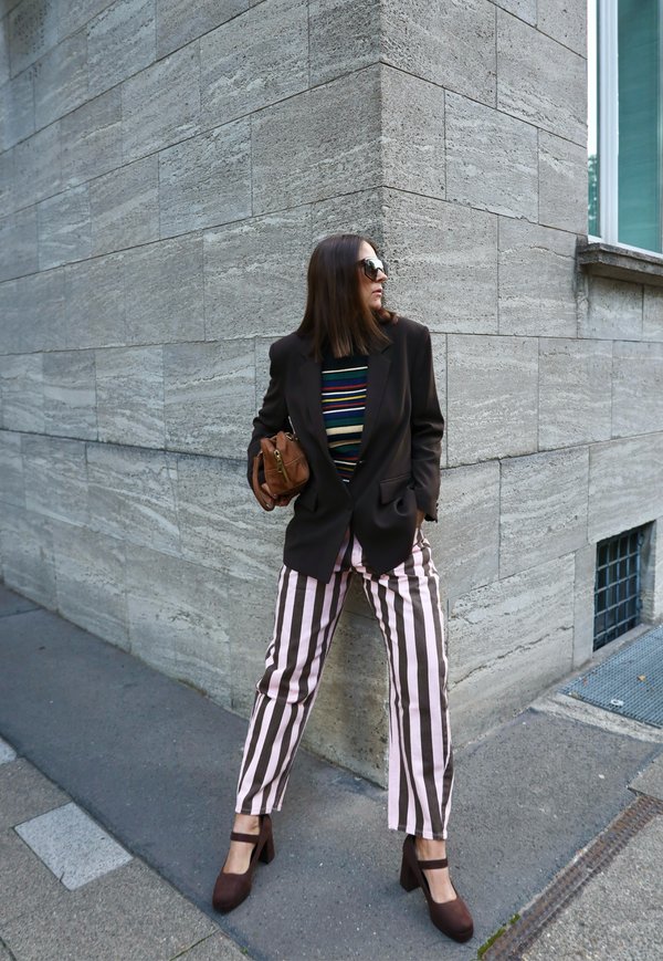 Mujer con blazer marrón, pantalones de rayas y tacones altos, de pie contra la esquina de una pared de piedra, sosteniendo un pequeño bolso marrón y llevando gafas de sol.