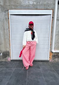 Suéter branco fofinho, calças largas xadrez vermelhas e brancas, boné vermelho, cachecol estampado e ténis cor-de-rosa. O fundo apresenta uma persiana rolô fechada.