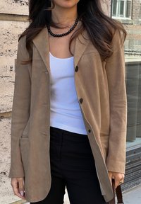 Ανοιχτό καφέ σουέτ μπουφάν με γιακά λαπέλ, κλείσιμο με έναν μόνο κουμπί και μπροστινή τσέπη, συνδυασμένο με λευκή ριχτή μπλούζα και μαύρο παντελόνι.