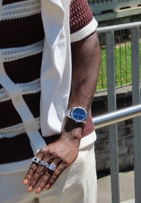 Ασημένιο ρολόι χειρός με μπλε καντράν, φορεμένο σε χέρι με τρία ασημένια δαχτυλίδια. Το φόντο περιλαμβάνει μια κουπαστή και πράσινη βλάστηση.