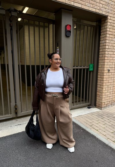 Chaqueta de cuero marrón de corte oversize, top blanco ajustado, pantalones de pierna ancha beige con cordón, bolso tejido negro y zapatos blancos.