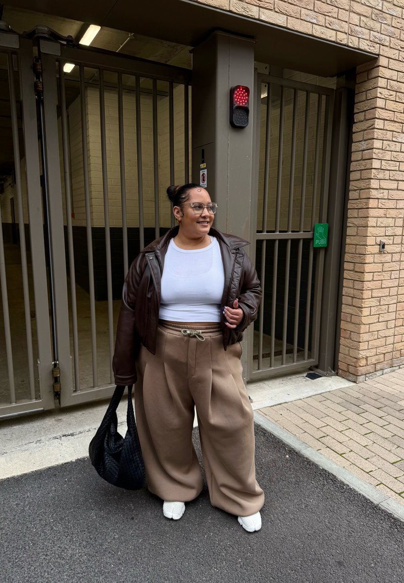 Brown leather oversized jacket, white fitted top, beige wide-leg trousers with drawstring, black woven handbag, and white shoes.