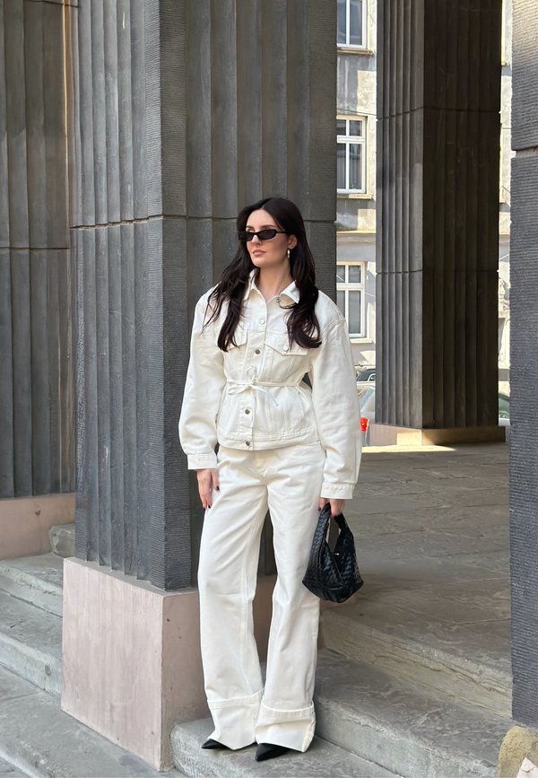 Kobieta ubrana w białą kurtkę i spodnie, w czarnych okularach przeciwsłonecznych, trzymająca czarną torebkę, stoi obok wysokich szarych kolumn na zewnątrz.