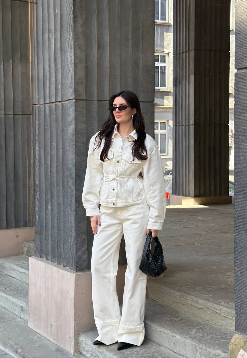 Donna che indossa giacca e pantaloni bianchi, occhiali da sole neri e tiene una borsa nera in mano, in piedi accanto ad alte colonne grigie all'aperto.