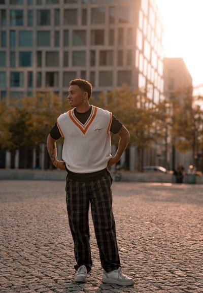Chaleco de punto blanco con detalles naranjas sobre una camiseta negra, combinado con pantalones de cuadros negros y zapatillas blancas. Entorno al aire libre con edificios.