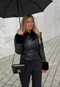 Veste en cuir noire avec poignets en fourrure, parapluie noir et petit sac à main noir avec ferrures dorées. Fond en carrelage gris.