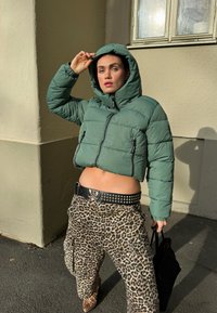 Izrezana, obložena zelena jakna s kapuljačom; crni remen s nitnama i kopčom; cargo hlače s leopardovim uzorkom; drži crnu torbu.