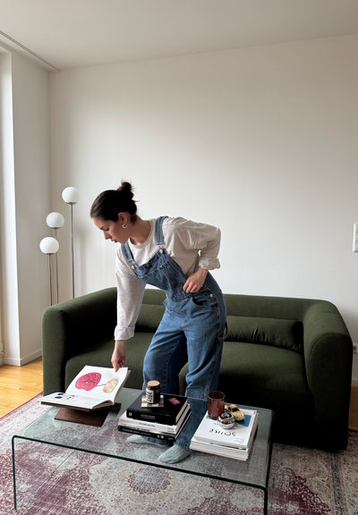 Mujer con peto vaquero mirando un libro abierto sobre una mesa de cristal en una sala de estar con un sofá verde y una lámpara de pie.
