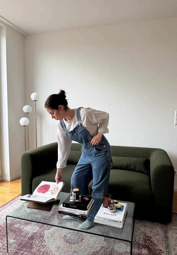 Vrouw in denim tuinbroek bladert in een open boek op een glazen salontafel in een woonkamer met een groene bank en een vloerlamp.