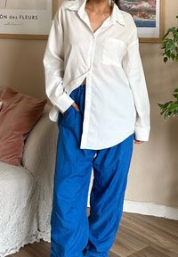 Λευκό πουκάμισο με κουμπιά και τσέπη, συνδυασμένο με χαλαρούς, υφασμάτινους μπλε παντελόνες. Το σύνολο έχει μια casual, χαλαρή σιλουέτα.
