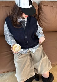 Personne portant une chemise rayée, un gilet marine, une cravate, une casquette de baseball et un short beige assise sur un canapé en cuir marron tenant une tasse de latte avec art.