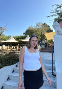Weißes Tanktop mit blauen Akzenten, schwarzer Rock und silberne Armbänder; steht auf Steinstufen. Im Hintergrund sind Zelte und Sitzmöglichkeiten im Freien zu sehen.