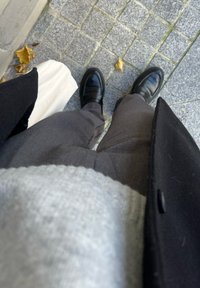 Cilvēks, kurš ģērbts melnā mētelī, pelēkās biksēs un melnās kurpēs, tur baltu somu, stāv uz pelēkām flīzēm, kurām apkārt izkaisītas dzeltenas lapas.