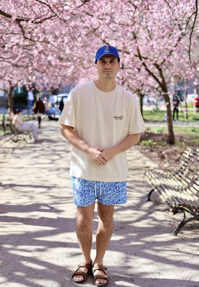 Camiseta de algodón beige con el texto "ORIGINALS", pantalones cortos de baño azules con estampado floral y sandalias marrones. El fondo presenta árboles en flor de color rosa.