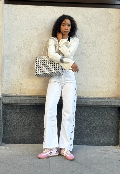Mujer con cabello rizado que lleva pantalones acampanados blancos con ojales, una blusa de manga larga color crema, zapatillas rosas y blancas, sosteniendo un bolso estampado.