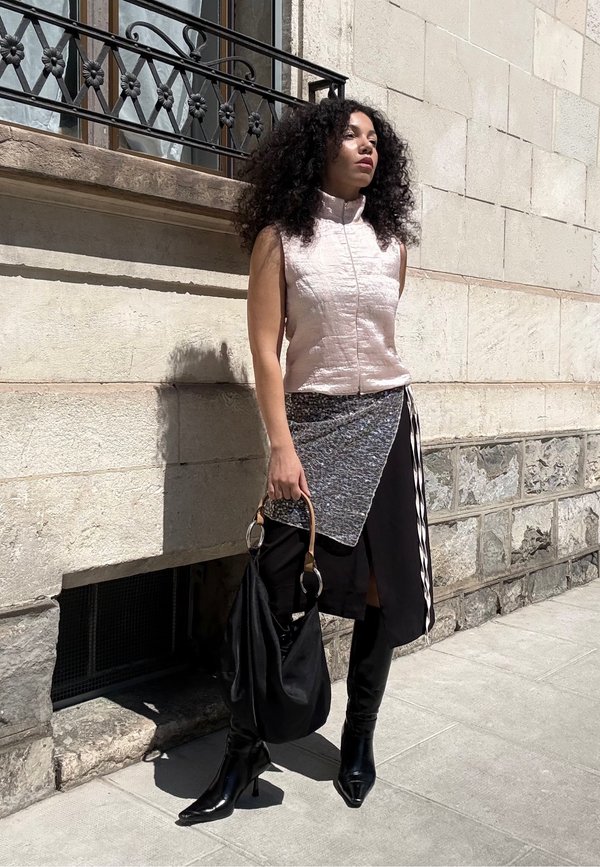 Donna con capelli ricci in piedi accanto a un muro di pietra, indossa un top leggero senza maniche, gonna asimmetrica, stivali neri e tiene una borsa nera.