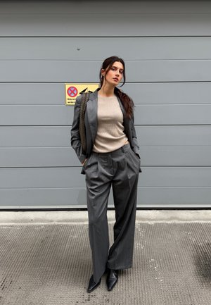 Traje gris a rayas con pantalones de pierna ancha y blazer ajustado, superpuesto a una blusa de punto beige con relieve. Botines negros de punta. Fondo texturizado.