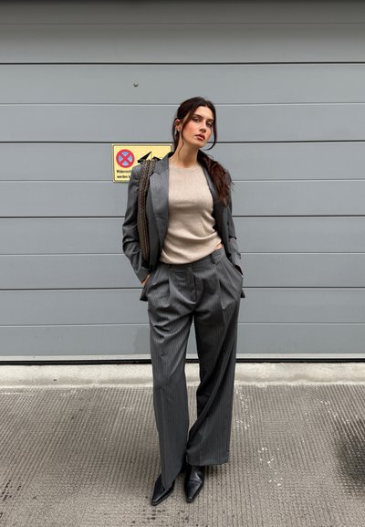 Traje gris a rayas con pantalones de pierna ancha y blazer ajustado, superpuesto a una blusa de punto beige con relieve. Botines negros de punta. Fondo texturizado.